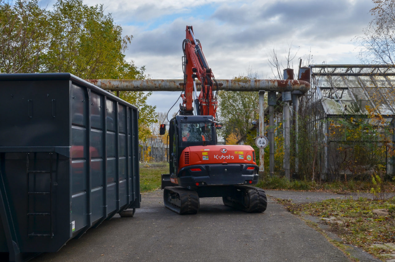 Neben einem Baucontainer und verfallenen Gewächshäusern steht ein Bagger. Auf dem Gelände einer ehemaligen Großgärtnerei beginnen in Kürze Abrissarbeiten.