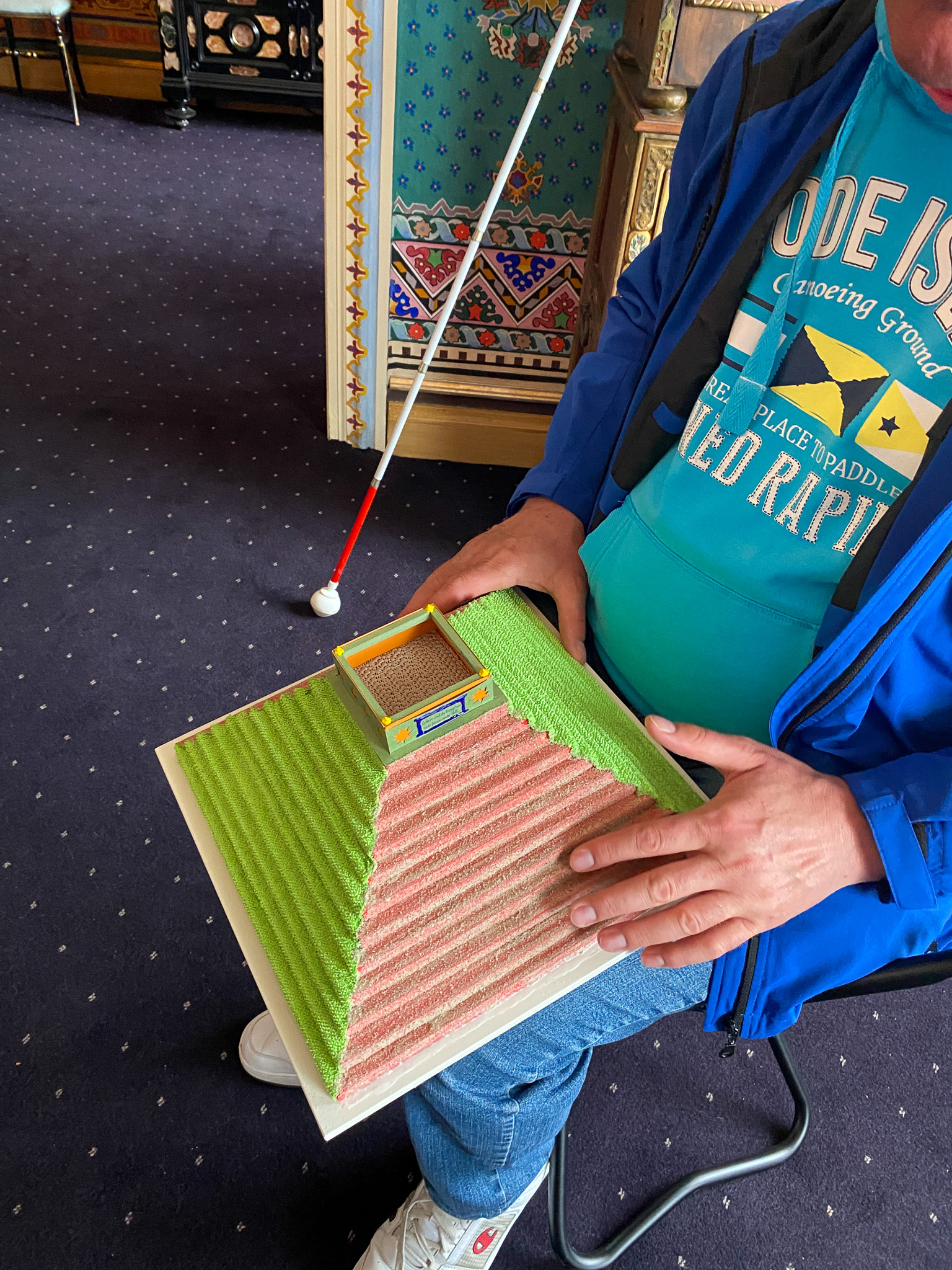 Ein Mann berührt und ertastet mit den Händen ein Tastmodell der Landpyramide. Die Pyramide hat mehrere Stufen und ein rechteckiges Geländer an der Spitze.