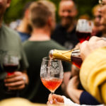 Eine Hand schenkt aus einer Weinflasche Rosewein in ein Weinglas. Im Hintergrund sind verschwommen Personen zu sehen.
