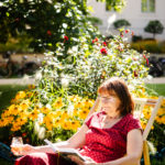 Eine Frau sitzt vor einem Blumenbeet mit einem Glas Weißwein und einem Buch in den Händen entspannt auf einem Liegestuhl.