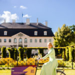 Eine Grau in einem langen grünen Kleid sitzt auf einem Lastenrad. Im Hintergrund ist Schloss Branitz zu sehen.