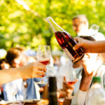 Eine Hand hält eine Flasche mit Rotwein zum Einschenken. Eine andere Hand ein halb gefülltes Weinglas. Im Hintergrund sind undeutlich Menschen zu erkenen.