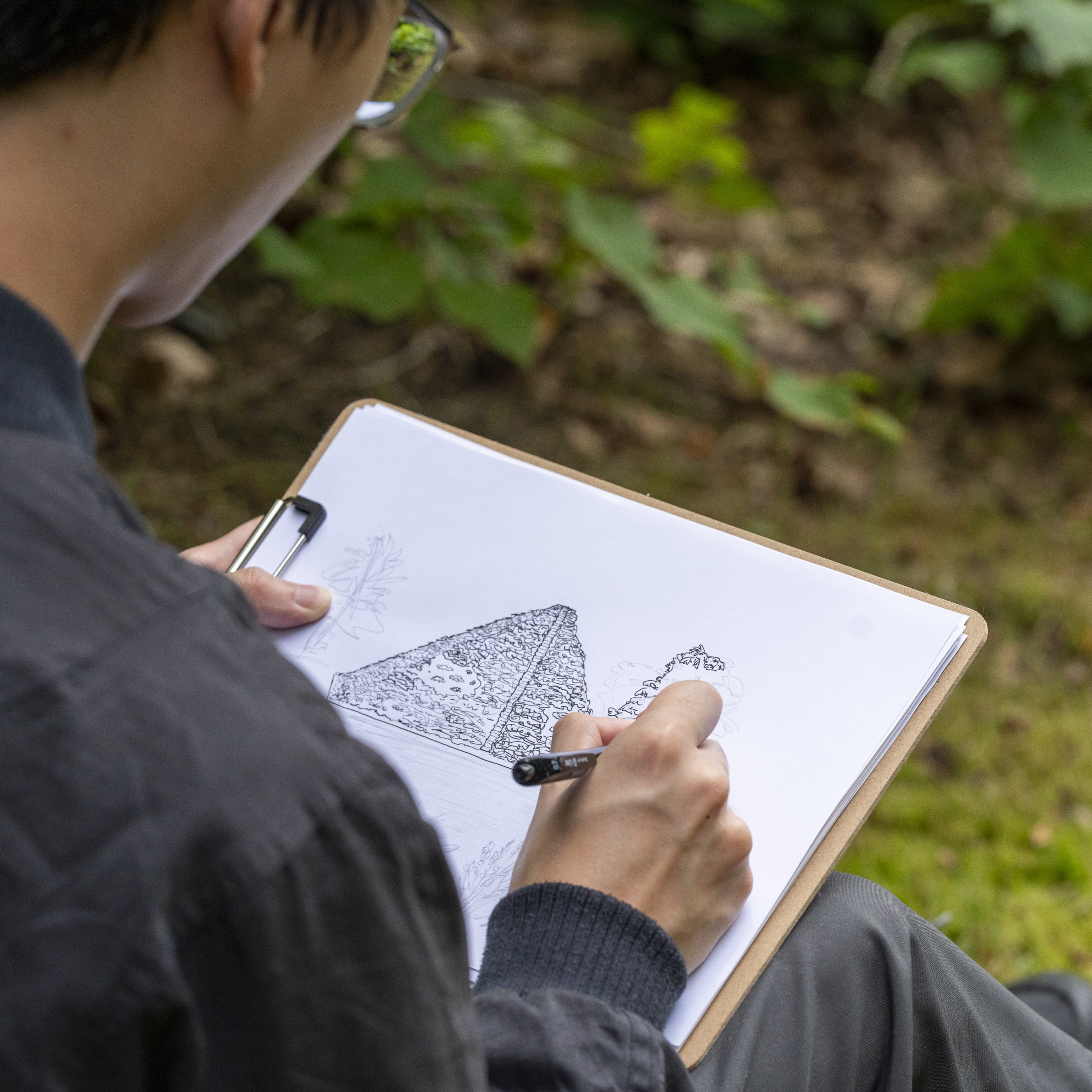 Ein junger Mann zeichnet mit Bleistift auf einem Papier, das an einem Klemmbrett befestigt ist, die Seeepyramide im Branitzer Park