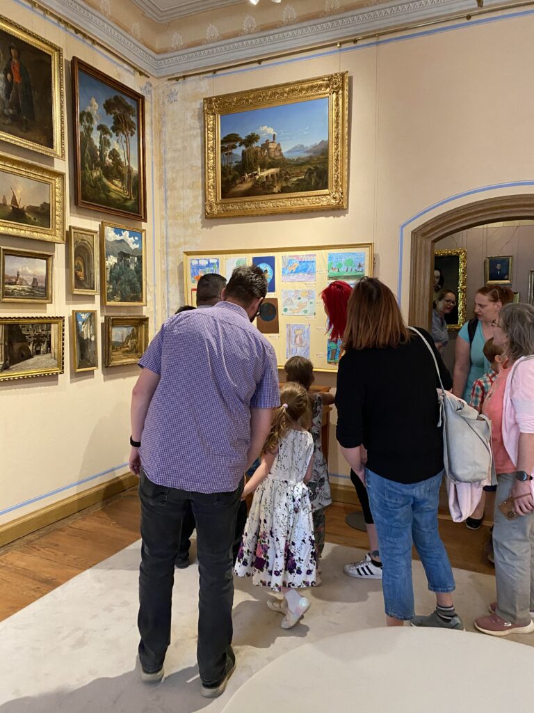 Familien mit Kindern betrachten Kinderzeichnungen, die in einer Ausstellung hängen.