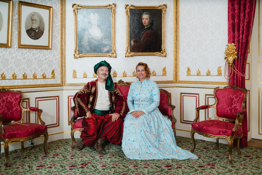 Zwei als Fürst und Fürstin gekleidete Personen sitzen auf einem Sitzmöbel in einem Schlosszimmer.