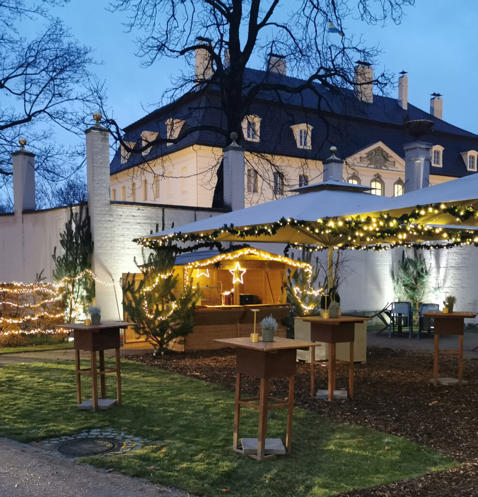 Schirme, Stehtische und eine Holzhütte mit Weihnachtsbeleuchtung in einem Hof. Im Hintergrund ist das Schloss Branitz.